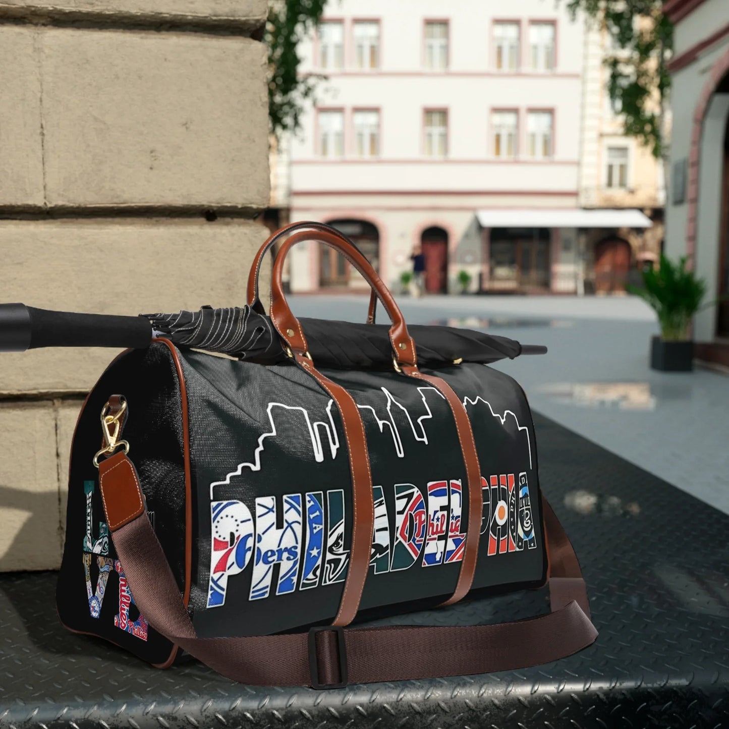Black Philadelphia-themed travel duffel bag with brown leather handles and folded umbrella, urban background